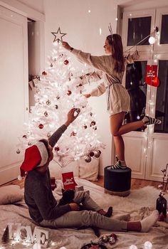 two people decorating a white christmas tree in a living room with lights and presents on the floor