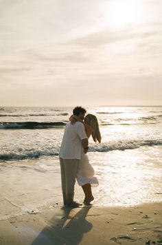 a man and woman hug on the beach as the sun sets behind them in the distance