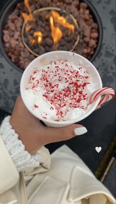 a person holding up a cup with whipped cream and candy canes in front of a fire pit