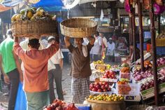 two men carrying baskets on their heads in front of a fruit and veggie market