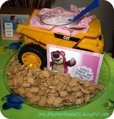 a table topped with a yellow toy dump truck filled with food next to a card