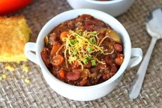 two white bowls filled with chili and cheese next to cornbread on a tablecloth