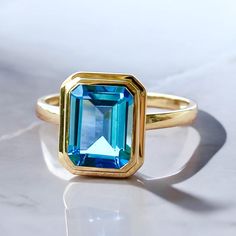 💎 14K Solid Gold Natural Swiss Blue Topaz Emerald-Cut Ring *Metal: 14K Solid Gold *Gemstone:    - **Topaz     - Cut: Emerald-Cut      - Color: Swiss Blue      - Clarity: Eye-Clean      - Size: 7 x 9 mm      - Total Carat Weight (CTW): 2.50 💓 Why You'll Love It:  The 14K Solid Gold Natural Swiss Blue Topaz Emerald-Cut Ring is a striking blend of vibrant color and timeless sophistication. At its center, a stunning emerald-cut Swiss blue topaz, measuring 8 x 10 mm, radiates a captivating blue tha Yellow Gold Blue Topaz Baguette Cut Ring, Blue Topaz Baguette Cut Ring In Yellow Gold, Yellow Gold Baguette Cut Blue Topaz Ring, Baguette Cut Blue Topaz Ring In Yellow Gold, Faceted Blue Topaz Ring In Fine Jewelry Style, Blue Emerald Ring With Bezel Setting, Emerald-cut Topaz Birthstone Ring In Yellow Gold, Emerald Cut Yellow Gold Topaz Birthstone Ring, Blue Topaz Ring With Emerald Cut And Bezel Setting