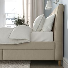 a bed with pillows and blankets on it in front of a window next to a potted plant