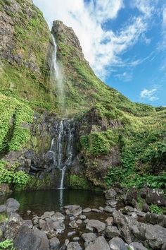 the waterfall is surrounded by rocks and greenery, with a blue sky in the background