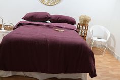 a bed with purple sheets and pillows in a room next to a white chair, table and clock on the wall