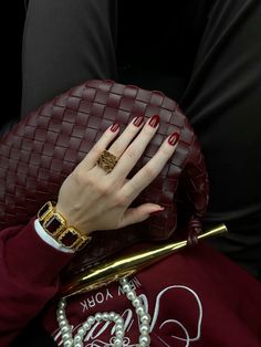 a woman's hand with red nails holding a burgundy purse and pearls on her wrist