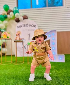 a little boy in a safari outfit standing on the grass with balloons and decorations behind him