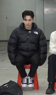 two young men sitting on red chairs in an airport terminal, one wearing a black jacket and the other white sweater