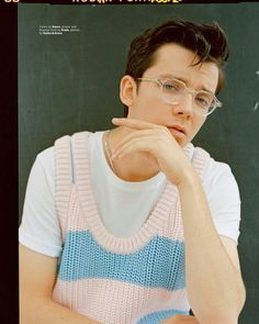 a young man wearing glasses sitting in front of a chalkboard with his hand on his chin