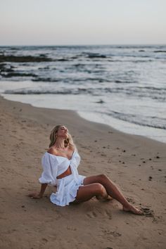 a woman sitting on top of a sandy beach next to the ocean wearing a white dress