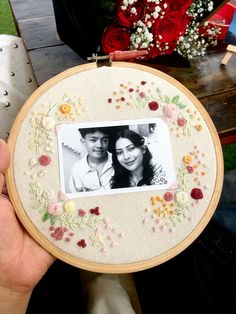 a man and woman are holding up an embroidered picture frame with flowers on it, in front of a bouquet of red roses