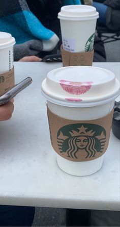 two cups of starbucks coffee on a table with people in the background looking at their cell phones