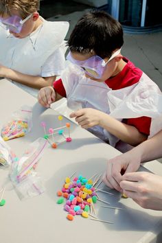 two boys in white lab coats and goggles working on crafting with colorful beads