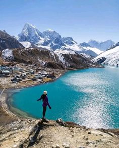 a person standing on top of a mountain next to a body of water with snow covered mountains in the background