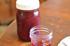 a jar of jam sitting on top of a wooden table next to a glass filled with liquid