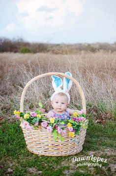 a baby wearing bunny ears sitting in a basket with flowers on the ground and grass