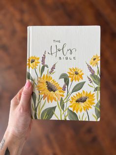 a person holding up a bible with sunflowers and lavender flowers on the cover