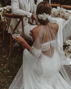 a bride and groom sitting at a table with flowers in their hair, holding wine glasses