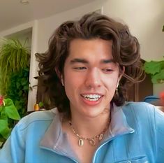 a young man with curly hair wearing a blue shirt and gold necklace smiling at the camera