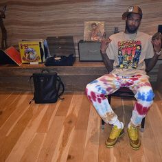 a man sitting on top of a wooden bench wearing tie dye pants and a hat