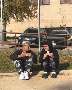 two people sitting on the ground eating food next to a parking lot with cars in the background