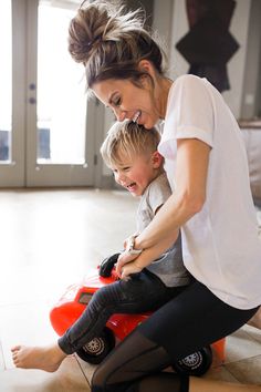 a woman sitting on top of a red toy car next to a little boy in black pants