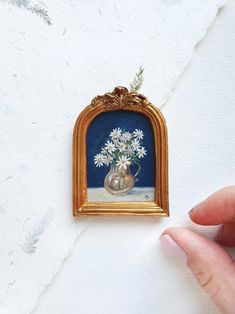 a hand is holding a miniature painting with daisies in a vase on the wall