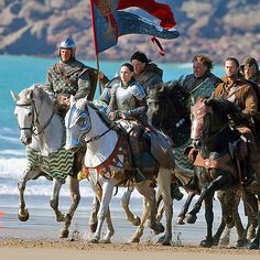 several people riding horses on the beach with a flag in front of them and one person holding a flag