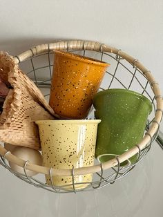 four different colored cups in a basket on top of a glass table with a white wall behind them