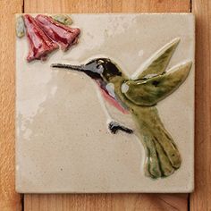 a ceramic tile with a hummingbird and flower on it's back side, sitting on a wooden surface