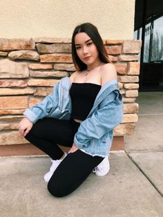 a woman sitting on the ground in front of a stone wall wearing black pants and a denim jacket