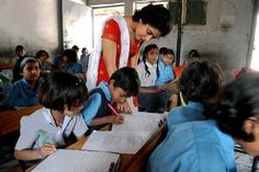 a woman standing in front of a group of children sitting at desks writing on paper