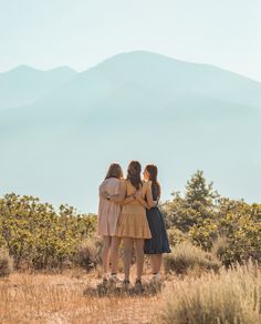 three girls standing in the middle of a field with their arms around each other and mountains in the background