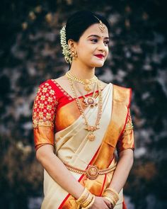 a woman in a white and red sari with gold jewelry on her neck, standing next to a stone wall