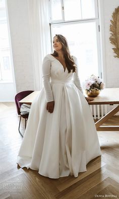 a woman standing in front of a window wearing a long white wedding dress with pockets