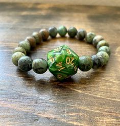 a green and gold beaded bracelet with a dice on it sitting on a wooden table