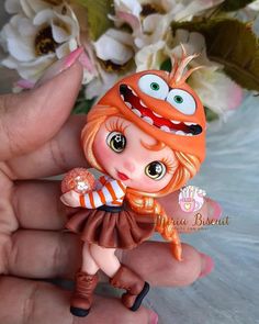 a hand holding a small doll with big eyes and an orange hat on it's head