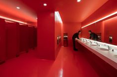 two men are standing in the bathroom with red walls and sinks, while one man is looking at himself in the mirror