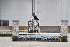 a person on a horse jumping over an obstacle with blue flowers in the foreground