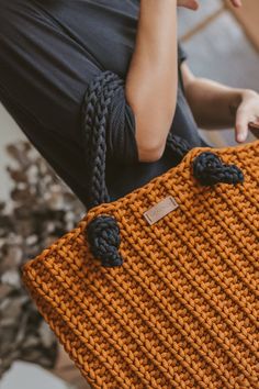 a woman is holding an orange knitted bag with black handles and tassels