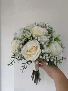 a hand holding a bouquet of white roses and greenery in front of a wall