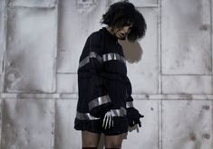 a woman with black hair and gloves standing in front of a white wall wearing an overstuffed sweater