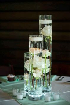 three tall vases filled with white flowers on top of a table next to candles