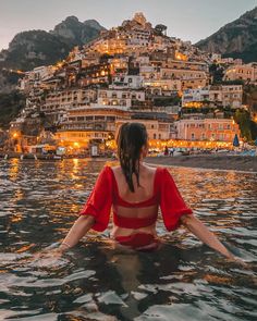 a woman is sitting in the water with her arms spread out and looking at buildings on top of a hill