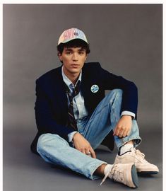 a young man sitting on the ground wearing a hat and tie with his hands in his pockets