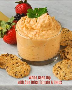 white bean dip with sun dried tomato and herbs in a glass bowl surrounded by crackers and strawberries