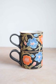 two black and orange floral mugs sitting on top of a wooden table next to each other