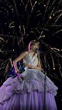 a woman in a purple dress holding a guitar and singing into a microphone with fireworks behind her