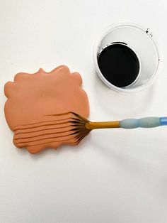 a cup of coffee next to a paintbrush on a white surface with an orange flower